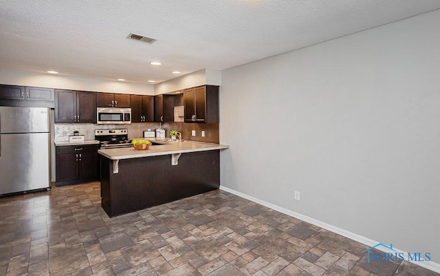 kitchen with appliances with stainless steel finishes, backsplash, dark brown cabinets, a kitchen bar, and kitchen peninsula