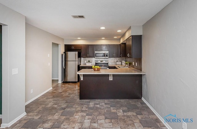 kitchen with a kitchen bar, decorative backsplash, kitchen peninsula, stainless steel appliances, and dark brown cabinets