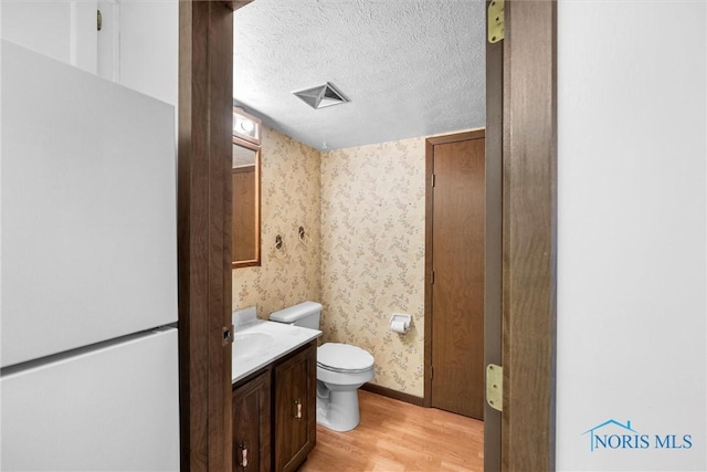 half bathroom with wallpapered walls, toilet, wood finished floors, a textured ceiling, and vanity