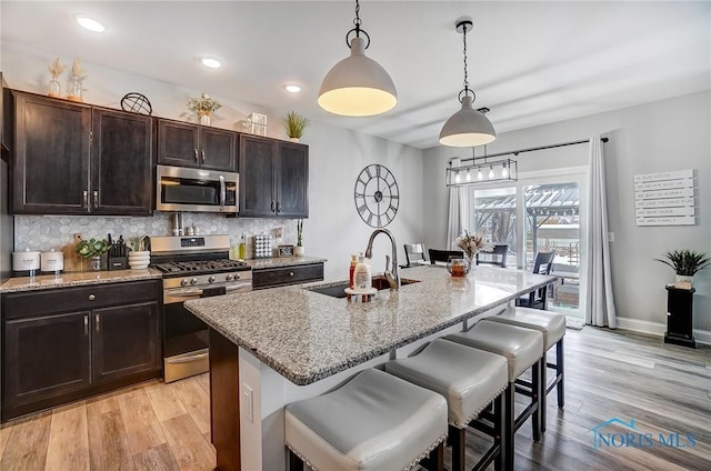 kitchen featuring appliances with stainless steel finishes, an island with sink, sink, hanging light fixtures, and light stone countertops