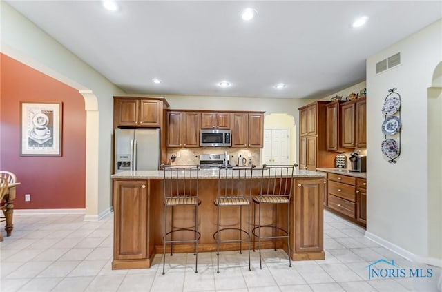 kitchen with backsplash, a center island with sink, stainless steel appliances, a kitchen breakfast bar, and light tile patterned flooring