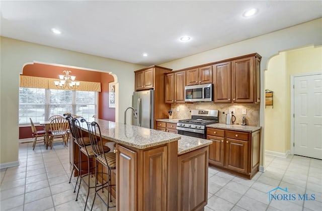 kitchen with tasteful backsplash, an inviting chandelier, appliances with stainless steel finishes, an island with sink, and light stone countertops