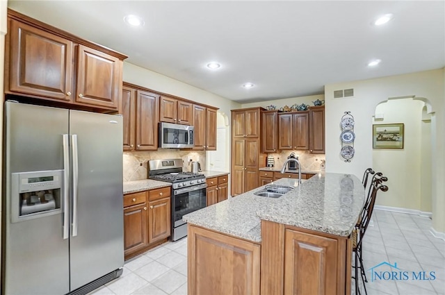 kitchen featuring sink, light stone counters, appliances with stainless steel finishes, a kitchen island with sink, and decorative backsplash