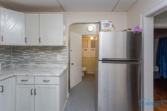 kitchen featuring light stone counters, a drop ceiling, stainless steel fridge, white cabinets, and backsplash