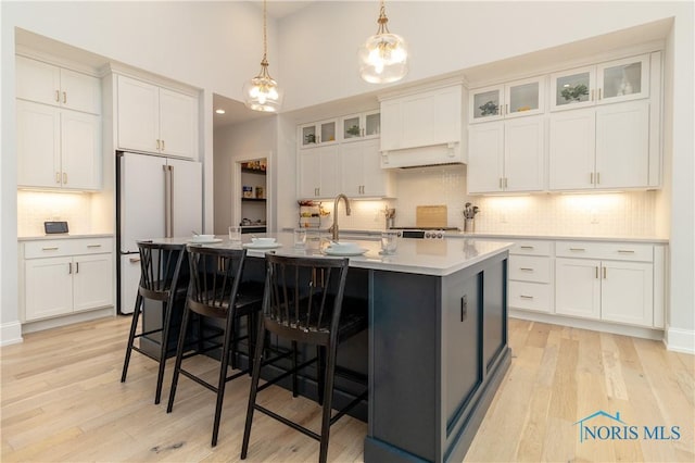 kitchen featuring high end white refrigerator, light hardwood / wood-style flooring, hanging light fixtures, a kitchen island with sink, and white cabinets