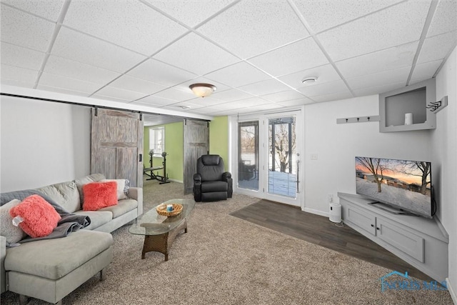 carpeted living area with a barn door, wood finished floors, a paneled ceiling, and baseboards