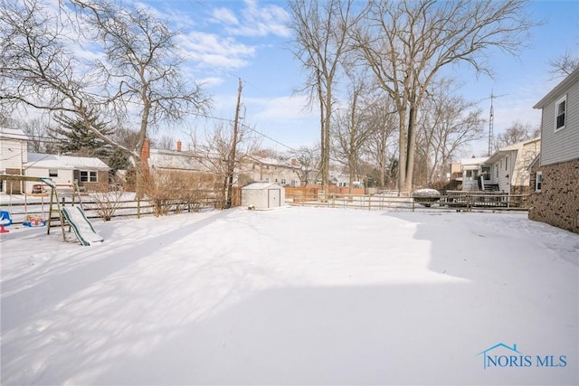 yard covered in snow featuring an outbuilding, a playground, a storage shed, fence, and a residential view