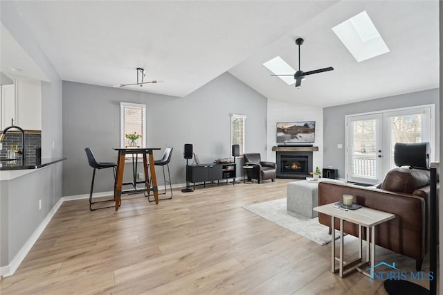 living area featuring light wood finished floors, baseboards, lofted ceiling with skylight, ceiling fan, and a lit fireplace