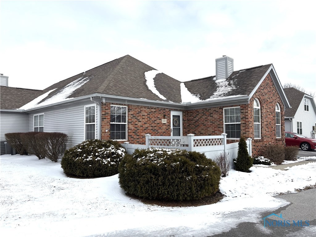 view of snow covered exterior featuring brick siding, a chimney, a shingled roof, and fence