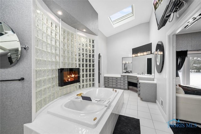 ensuite bathroom featuring vaulted ceiling with skylight, a glass covered fireplace, tile patterned floors, a garden tub, and vanity