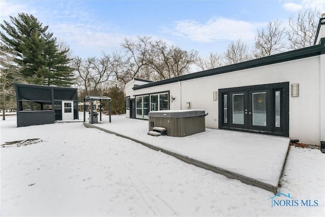 snow covered house with french doors, a hot tub, and stucco siding