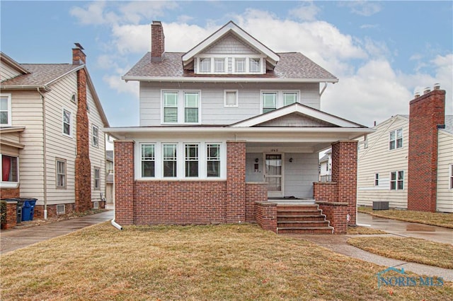 american foursquare style home featuring covered porch, brick siding, a front lawn, and roof with shingles