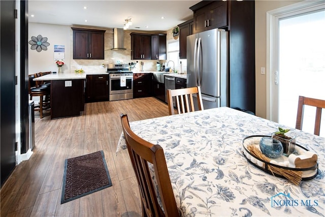 kitchen featuring stainless steel appliances, light countertops, dark brown cabinetry, wall chimney range hood, and a kitchen bar