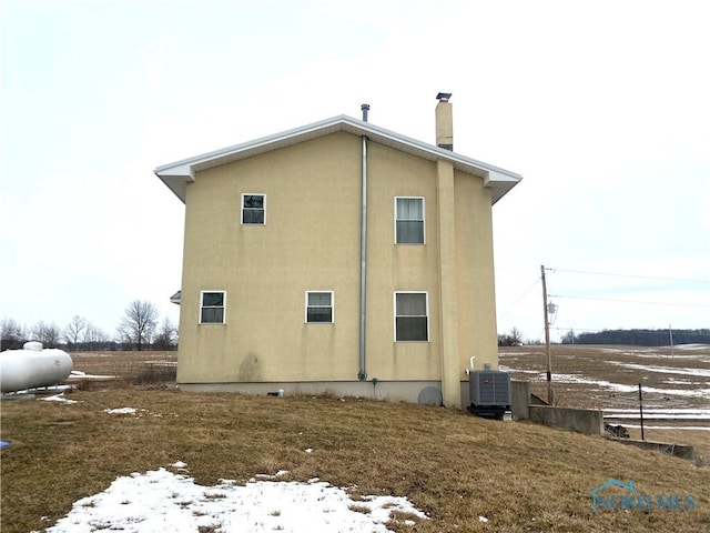 snow covered property with central air condition unit, a chimney, and stucco siding