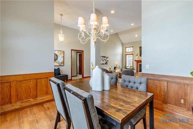 dining room featuring a wainscoted wall, a fireplace, a chandelier, high vaulted ceiling, and light wood-type flooring