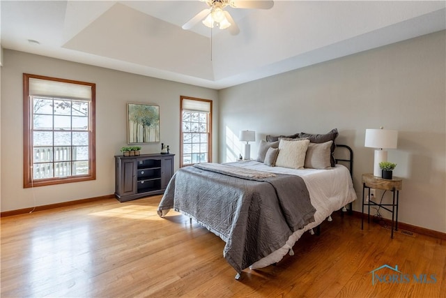 bedroom with a tray ceiling, light wood-type flooring, a ceiling fan, and baseboards