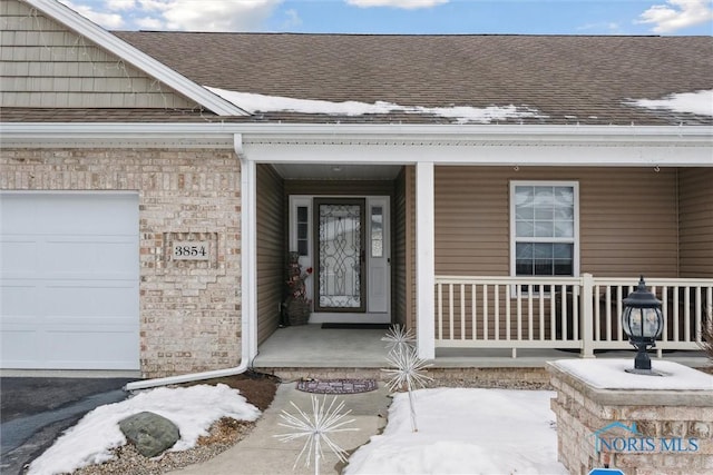 snow covered property entrance featuring an attached garage, a porch, a shingled roof, and brick siding