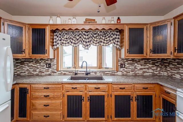 kitchen featuring a sink, backsplash, freestanding refrigerator, brown cabinets, and glass insert cabinets