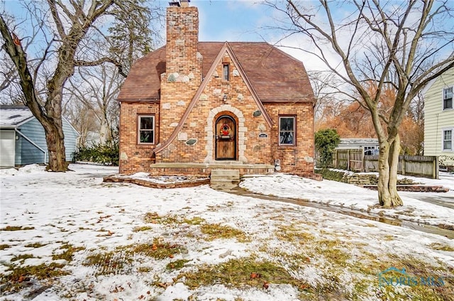 tudor house with a chimney, a detached garage, roof with shingles, fence, and brick siding