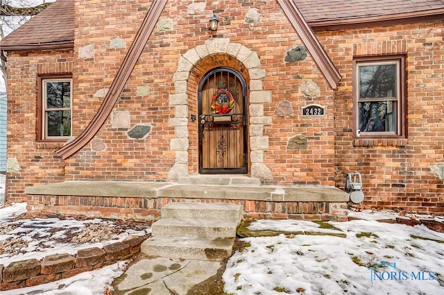 snow covered property entrance with a shingled roof and brick siding