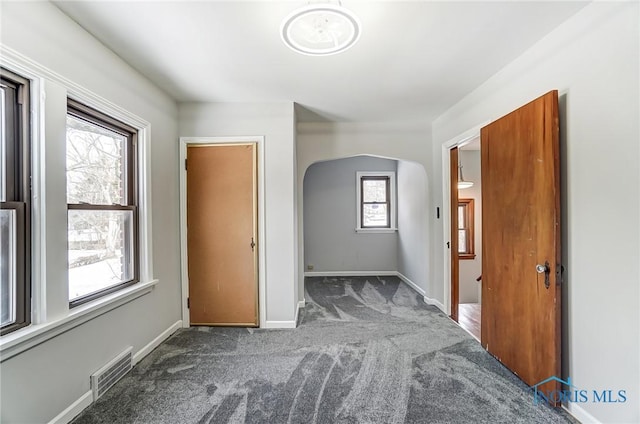 empty room with arched walkways, dark carpet, visible vents, and baseboards