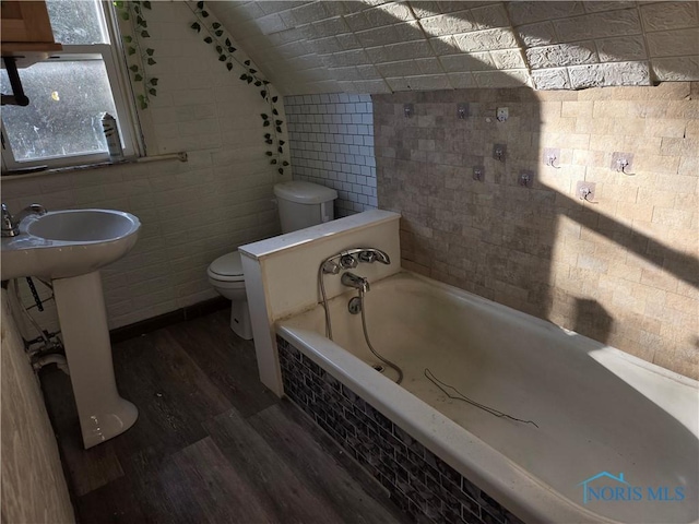 bathroom with a garden tub, toilet, wood finished floors, tile walls, and vaulted ceiling