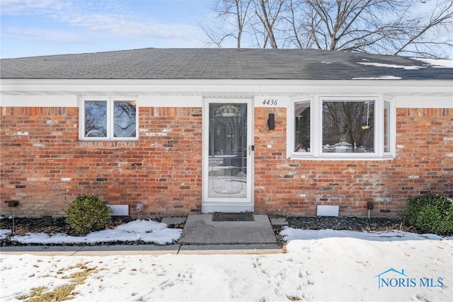 snow covered property entrance featuring crawl space, a shingled roof, and brick siding