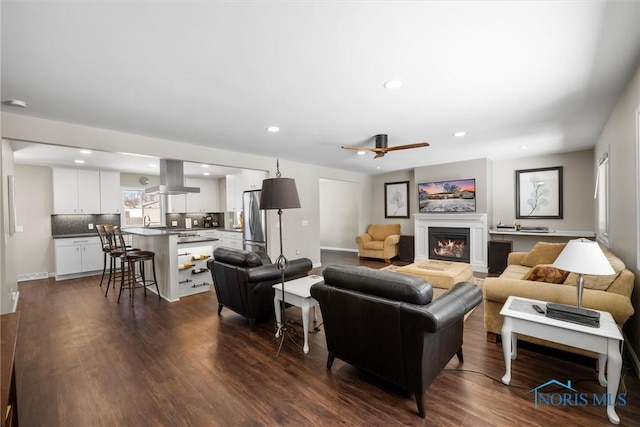 living room with dark wood-type flooring, recessed lighting, a warm lit fireplace, and ceiling fan