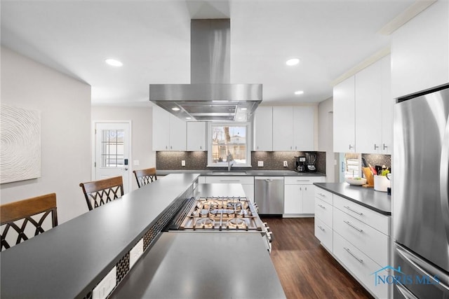 kitchen with stainless steel appliances, dark countertops, white cabinets, a sink, and island range hood