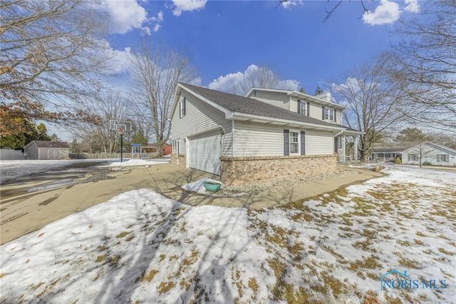 snow covered property featuring driveway, a storage shed, a chimney, an attached garage, and brick siding