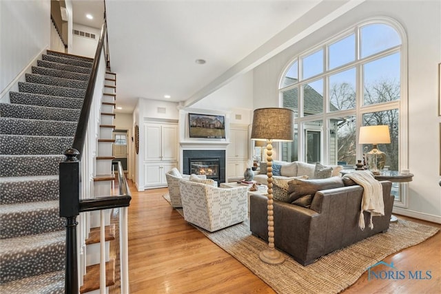 living area with visible vents, light wood-style flooring, a high ceiling, a glass covered fireplace, and stairs