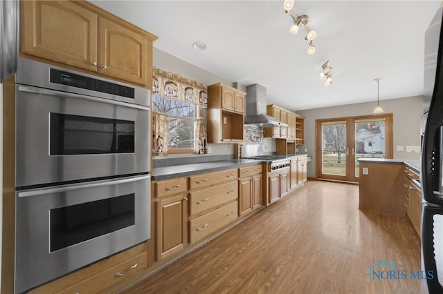 kitchen featuring light wood-style floors, hanging light fixtures, appliances with stainless steel finishes, wall chimney exhaust hood, and dark countertops