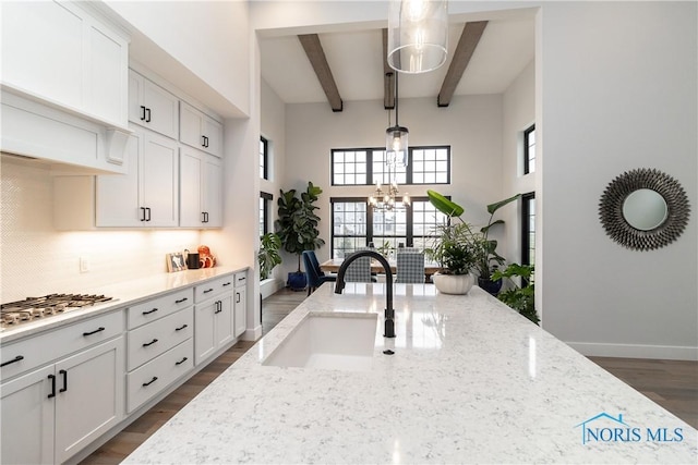 kitchen with stainless steel gas stovetop, decorative backsplash, a sink, light stone countertops, and beamed ceiling