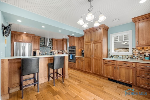 kitchen with brown cabinetry, a breakfast bar, freestanding refrigerator, double wall oven, and wall chimney range hood