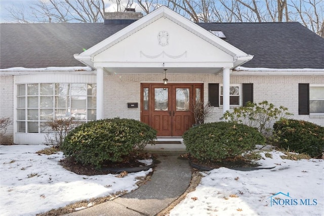 snow covered property entrance featuring roof with shingles, brick siding, and a chimney