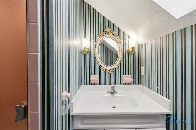 bathroom with a textured ceiling, vanity, and wallpapered walls