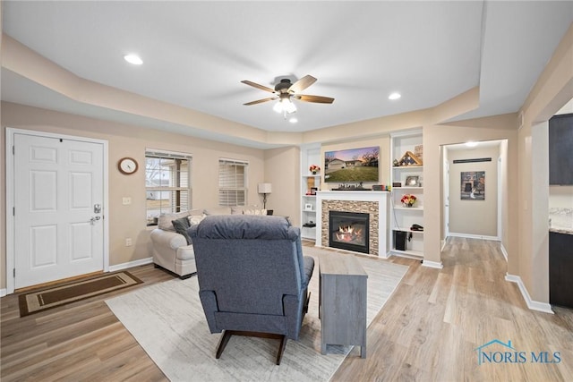 living area featuring ceiling fan, a stone fireplace, light wood finished floors, and baseboards