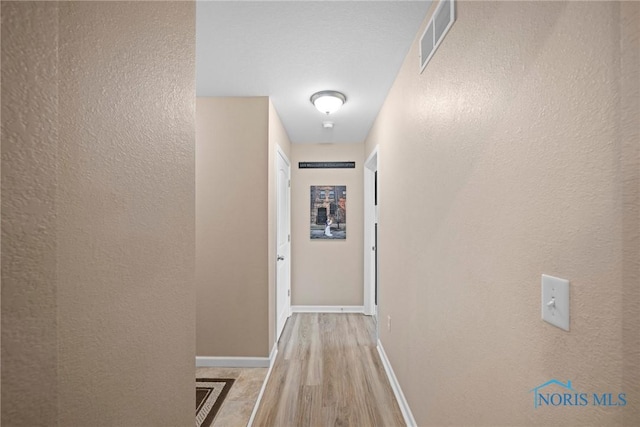 hallway featuring visible vents, a textured wall, baseboards, and wood finished floors