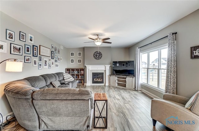 living room featuring a ceiling fan, light wood-type flooring, a glass covered fireplace, and baseboards