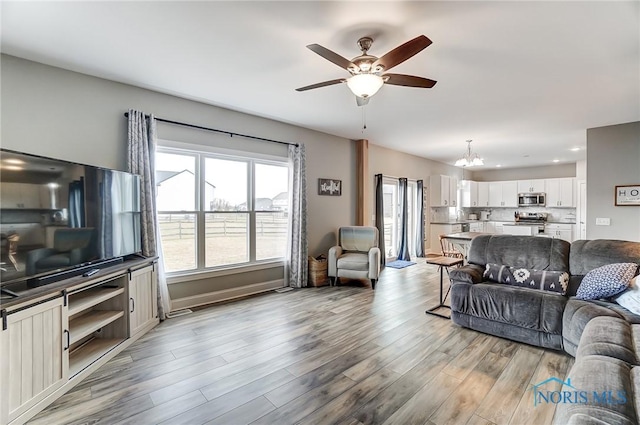 living area with ceiling fan with notable chandelier, light wood finished floors, and baseboards