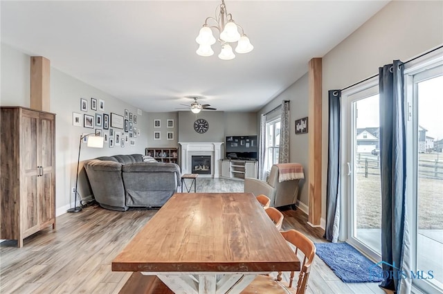 dining space with light wood-style flooring, ceiling fan with notable chandelier, baseboards, and a glass covered fireplace