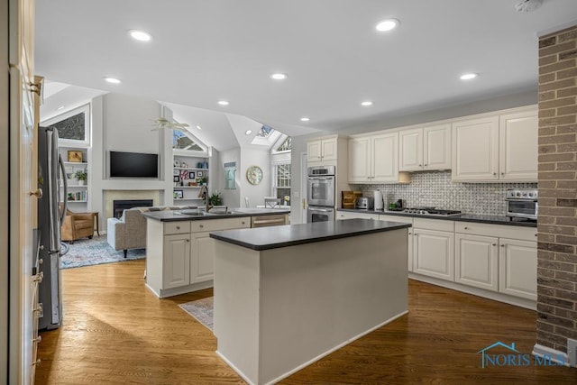 kitchen featuring a fireplace, dark countertops, appliances with stainless steel finishes, a kitchen island, and vaulted ceiling with skylight