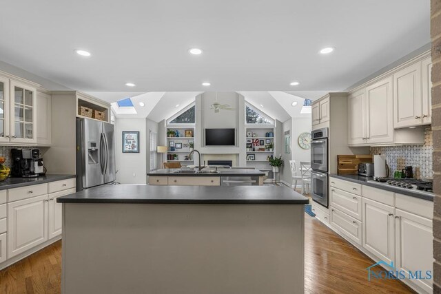 kitchen with stainless steel appliances, a sink, lofted ceiling with skylight, dark countertops, and an island with sink