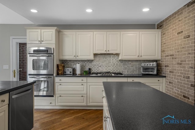 kitchen featuring tasteful backsplash, dark countertops, brick wall, appliances with stainless steel finishes, and wood finished floors