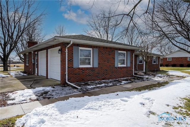 snow covered property featuring driveway, an attached garage, a chimney, and brick siding