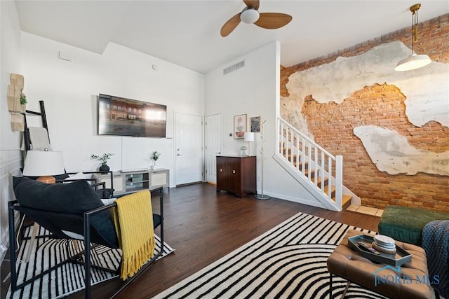 living room with ceiling fan, brick wall, wood finished floors, visible vents, and stairs