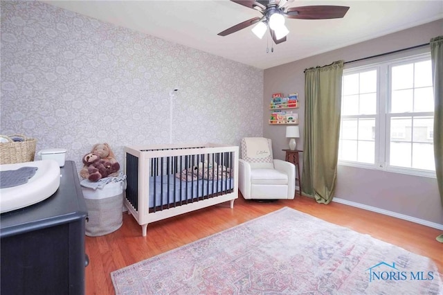 bedroom with a ceiling fan, wood finished floors, a crib, baseboards, and wallpapered walls