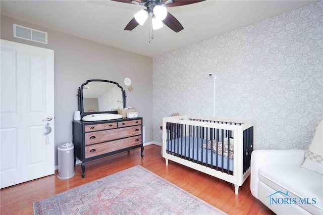 bedroom with wallpapered walls, visible vents, ceiling fan, wood finished floors, and a nursery area