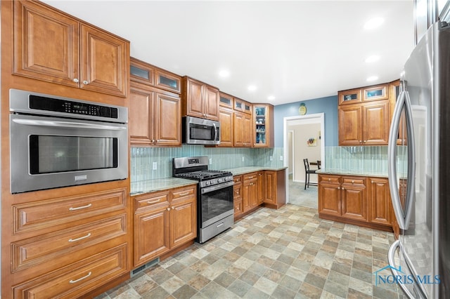 kitchen with light stone counters, stainless steel appliances, brown cabinets, decorative backsplash, and glass insert cabinets