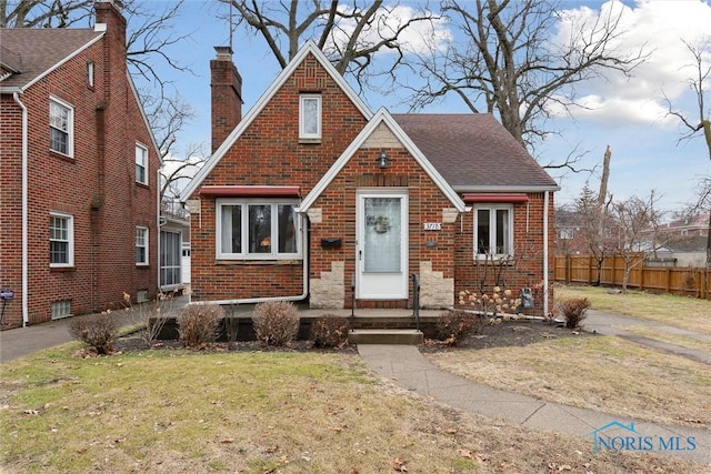 tudor home with brick siding, fence, roof with shingles, a chimney, and a front yard